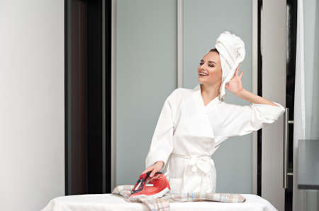 A young cheerful woman after a shower, wearing a white bathrobe and a towel on her head ironing clothes in the home interiorの写真素材