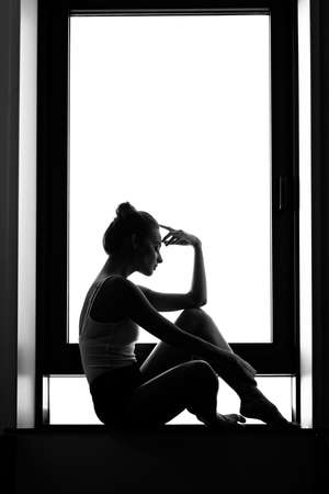 Silhouette of a woman sitting thinking on a window sill. Black and white photography. lonelinessの写真素材