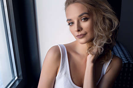 Close-up portrait of gorgeous brown-haired young woman. sitting next to the window. She touches her...