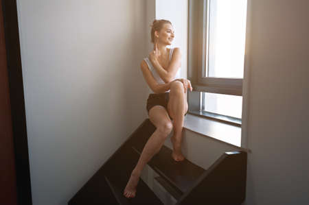 a slender girl with a brown woman sits on the steps on a bright terrace and looks out the window.の写真素材