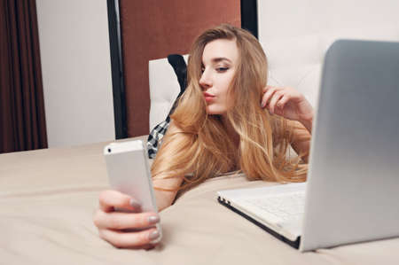 young blond hipster girl looking in the phone lying on the bed with laptop, writing letters,の写真素材