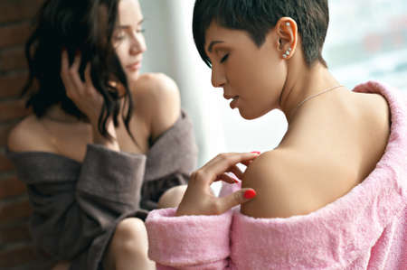 Two happy sexy young women, friends, in dressing gowns, sit at the window, on the floor, face to face. Unconventional couple.の写真素材