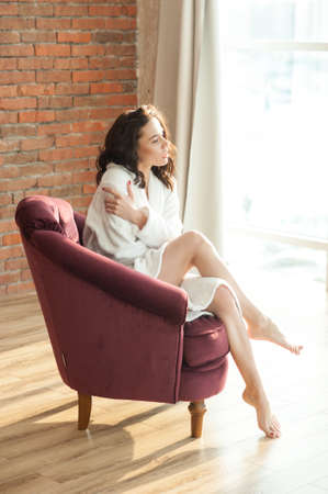 A young beautiful woman in a white terry dressing gown sits in a chair and looks out the window. Background, red brick wall.の写真素材