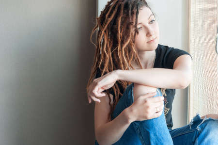 Portrait of a young girl in dreadlocks and torn jeans sitting on the window of the houseの写真素材