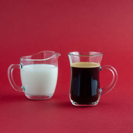 Transparent cup of black coffee and milk jug placed on a red surfaceの写真素材