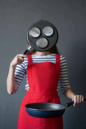 The image of a young woman in a cook apron covered her face with a frying pan for cooking fritters,...