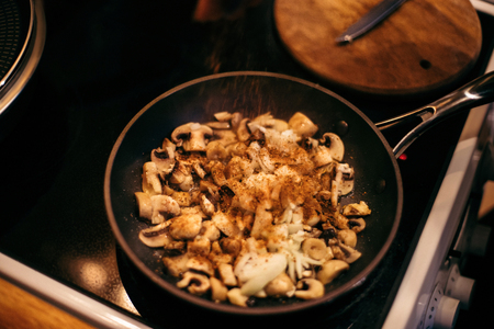 A woman is frying mushrooms and carrots in the kitchen. The process of frying in a frying pan. Preparation of food by prescription. Home kitchen. Mom cooks fried mushrooms and carrots.の写真素材