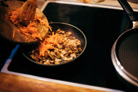 A woman is frying mushrooms and carrots in the kitchen. The process of frying in a frying pan. Preparation of food by prescription. Home kitchen. Mom cooks fried mushrooms and carrots.の写真素材
