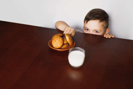 The child tries to steal oatmeal cookies and homemade milk from the table in the kitchen. A healthy morning breakfast.の写真素材