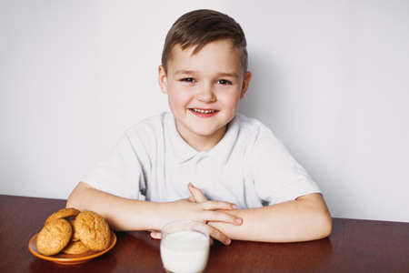 A young boy is sitting at a wooden table with oatmeal cookies and a glass of homemade milk. He smiled against the white background. Healthy food.の写真素材