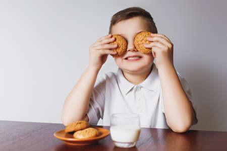 A young boy holds oatmeal cookies and closes his eyes. The boy is sitting in the kitchen with biscuits and milk.の写真素材
