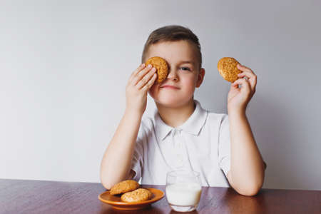 A young boy holds oatmeal cookies and closes his eyes. The boy is sitting in the kitchen with biscuits and milk.の写真素材