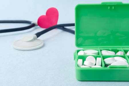 Green pillbox with white pills on the table. In the background, a stethoscope and a small red heart.