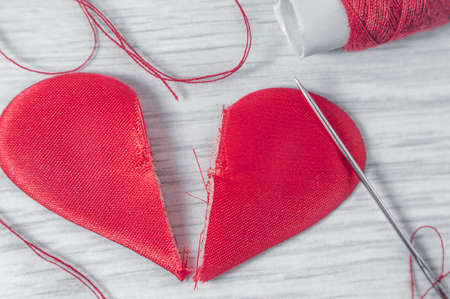 The heart is cut into two halves and lies on a white wooden table. Next to it is a needle with a threaded thread and a spool of red thread.の写真素材