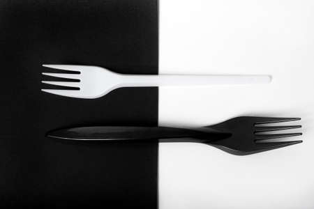 Two plastic forks, black and white, lie parallel to each other, the teeth pointing in different directions. The background is divided into two parts, black and white. Forks lie across the background.の写真素材