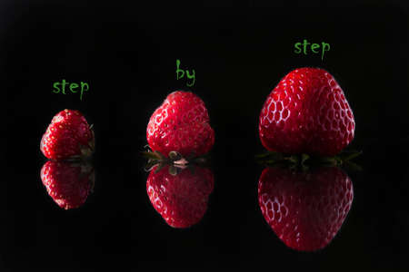 Concept of business development, career growth, motivational concept. On a black background, three red strawberries of different sizes. Above the strawberries is written in green letters: "step by step".の写真素材
