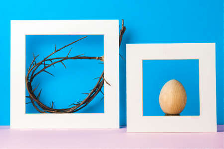 Still-life. In a white frame, a crown of thorns and a wooden egg. Blue background.
