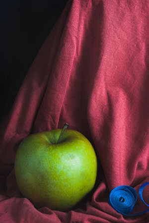 A green apple on a red cloth background, next to a blue measuring tape. The concept of proper nutrition and a healthy lifestyle.の写真素材