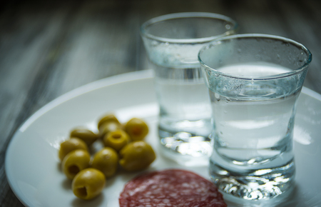 Two shots of vodka, olives and salami slices on white plateの写真素材