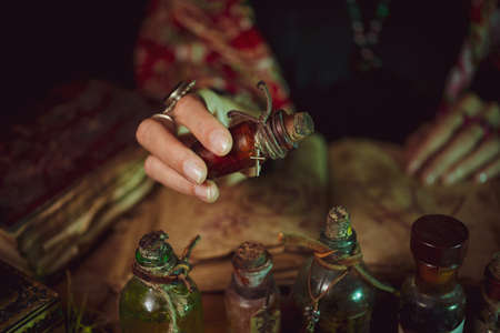 Female witch making potion on dark background, magic bottles with potions and candles on table of...