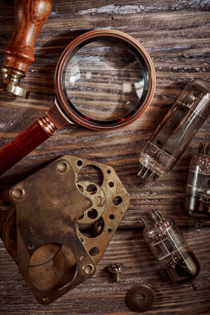 Steampunk still life - old vintage objects on a wooden background. Leather wrist watch, dark glasses, old tube lamps, magnifying glass, gearsの写真素材