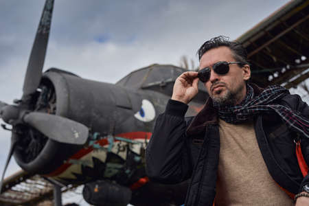 Oldschool pilot in the bomber jacket posing near the aircraft, aviator male with scarf and sunglassesの写真素材