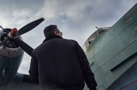 Oldschool pilot in the bomber jacket posing near the aircraft, aviator male with scarf and...