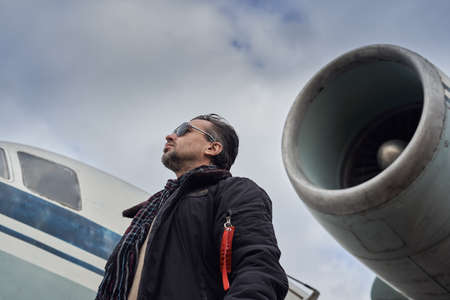 Oldschool pilot in the bomber jacket posing near the aircraft, aviator male with scarf and sunglassesの写真素材