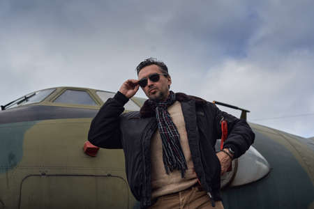 Oldschool pilot in the bomber jacket posing near the aircraft, aviator male with scarf and sunglassesの写真素材