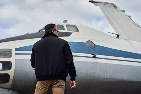 Oldschool pilot in the bomber jacket posing near the aircraft, aviator male with scarf and sunglassesの写真素材
