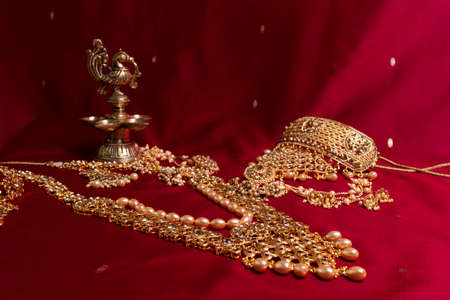 Indian traditional bronze oil lamp in the shape of a bird and gold Indian female jewelry on the red sari background. traditional Indian religious ceremony of Hindusの写真素材