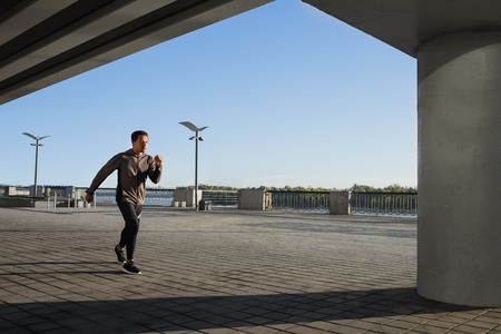 Young fit man in grey windbreaker running at outdoors, autumn running, attractive dark skinned runner jogging fast, sport conceptの写真素材