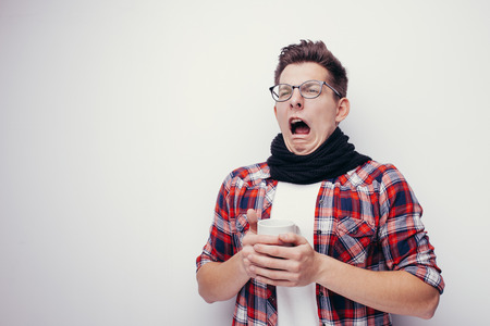 Man with flu and fever wrapped in scarf holding cup of healing tea isolated over white.の写真素材
