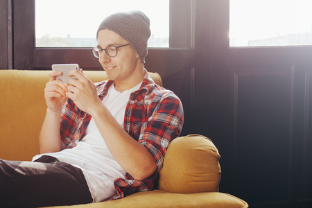 Beautiful young man relaxing on sofa and looking at his smart phoneの写真素材