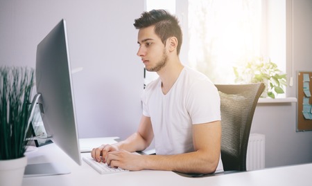 Young worker sitting in an office at the computer. Freelancer in a white shirt at work. The designer sits in front of a window in the workplace.の写真素材