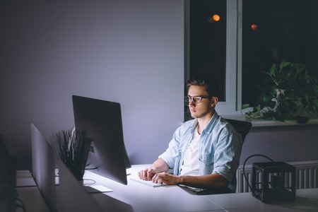 Young man working on computer at night in dark office. The designer works in the later time. A young man sits at the computerの写真素材