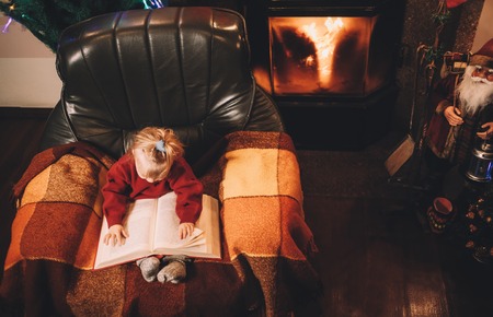 Little girl reading a bedtime story by the fireplace. Pre-christmas mood. New Years is soonの写真素材