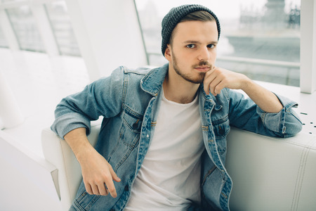 Young man is sitting on sofa and thinking about his life. he is looking at camera holding up his headの写真素材