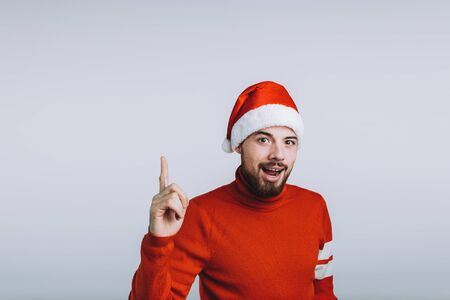 Winter concept - Christmas holiday. Happy young man is pointing up with his finger. He is wearing Santa's hat and red sweater. Isolated on white backgroundの写真素材