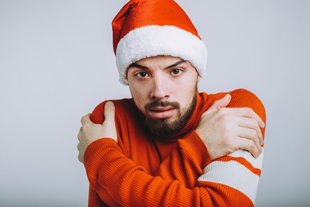 Handsome guy with beard hugging himself on white background. Serious man is looking at camera. He is wearing warm red sweater and santa claus hatの写真素材