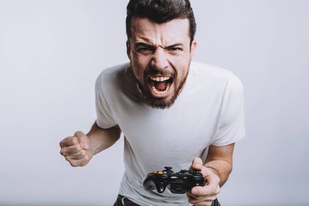 Young attractive guy with beard holding joystick. He is angry with his enemies because he lost the fight. He is screaming of fury and having fist. Isolated on white backgroundの写真素材