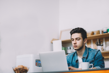 Young serious man with beard working on laptop. He is sitting and watching deep video. He is sittting in the kitchen with tea and cookies. Place for your text or logoの写真素材