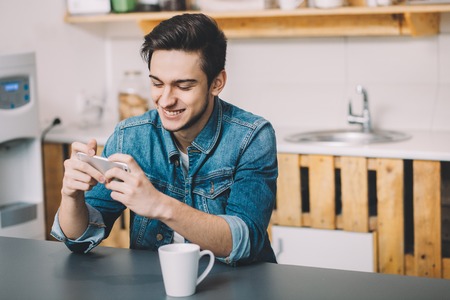 Young man sitting at a table in the kitchen with a cup of tea or coffee watching funny video. He is smiling because of seenの写真素材