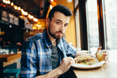 Young man having lunch at cafe. Serious but delighted handsome guy looking at camera holding cup with warm teaの写真素材