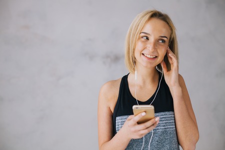 Beautiful young woman in sports wear and earphones is listening to music using a smartphone. Attractive blond girl is smiling and looking somewhere near. Isolated. Close upの写真素材