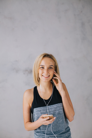 Beautiful young woman in sports wear and earphones is listening to music using a smartphone. Nice happy girl in grey top relaxing after work out. Isolated. Close upの写真素材