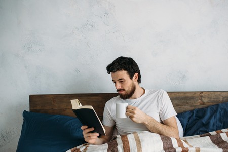 Bearded man reading a big book lying in his bedroom. Attractive smiling guy reeling off some story or interesting events holding book in one hand and cup of coffee in another one.の写真素材