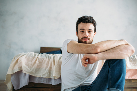 Young man sitting down on the floor near the bed. Attractive cute guy has folded his arms on knee and looking a camera. Close upの写真素材