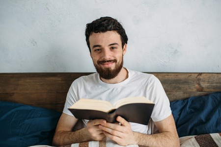 Bearded man reading a big book lying in his bedroom under the blanket and holding it with both hands. Smiling attractive guy perusing some story and looking right at the camera. Close upの写真素材