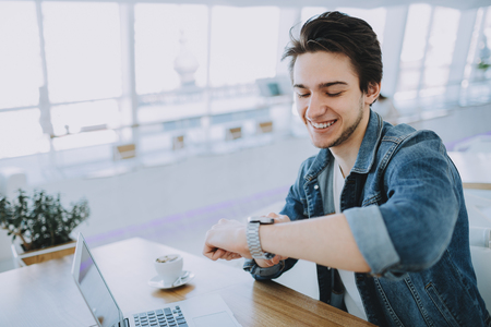 Young stylish man working on a macbook or laptop in cafe while having coffee and checking his nice watch. Happy guy in jeans jacket pointing at clock faceの写真素材